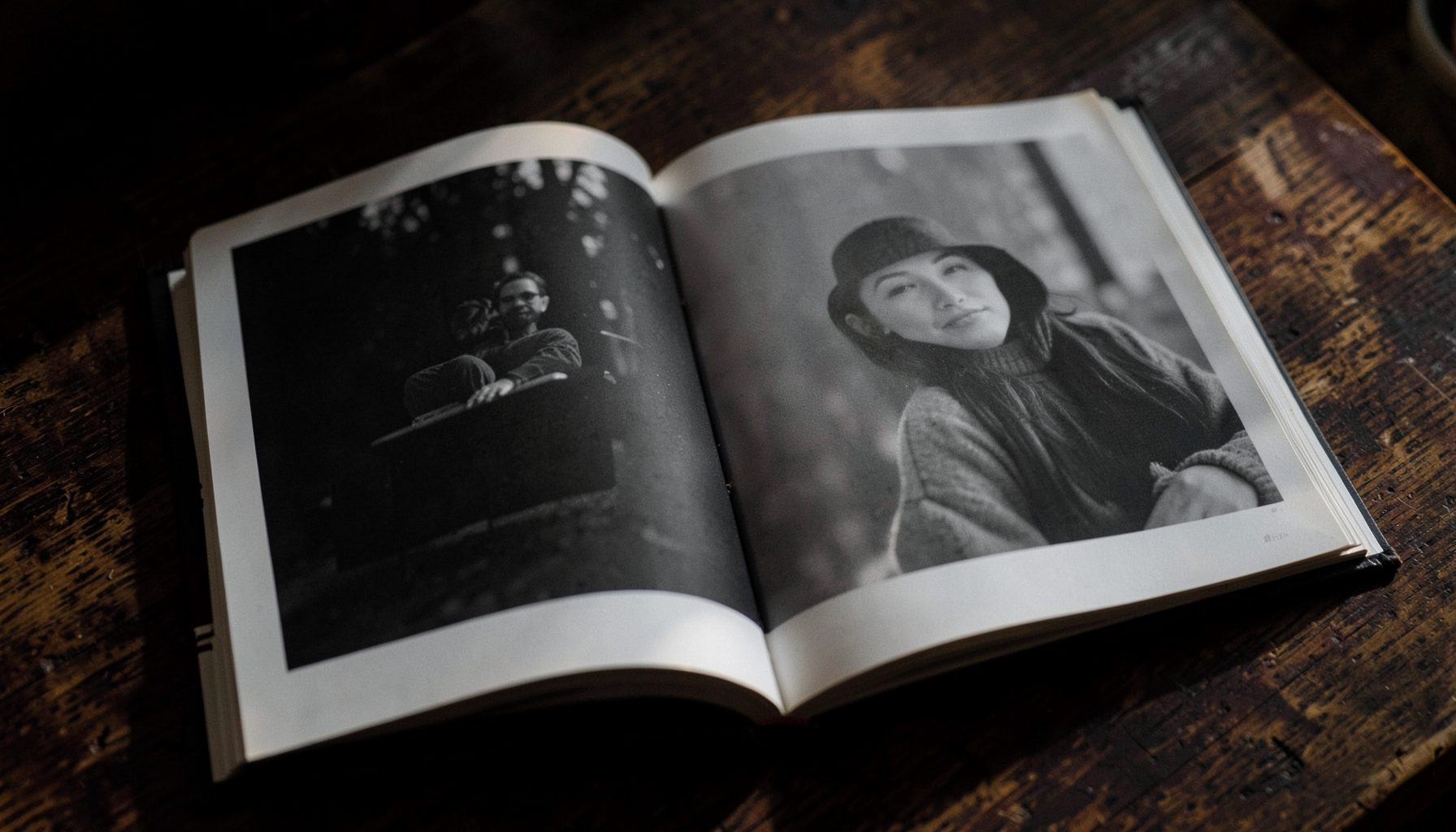 A moody black-and-white photography book open on a dark wooden table, soft natural light falling across the page, deep shadows and warm midtones