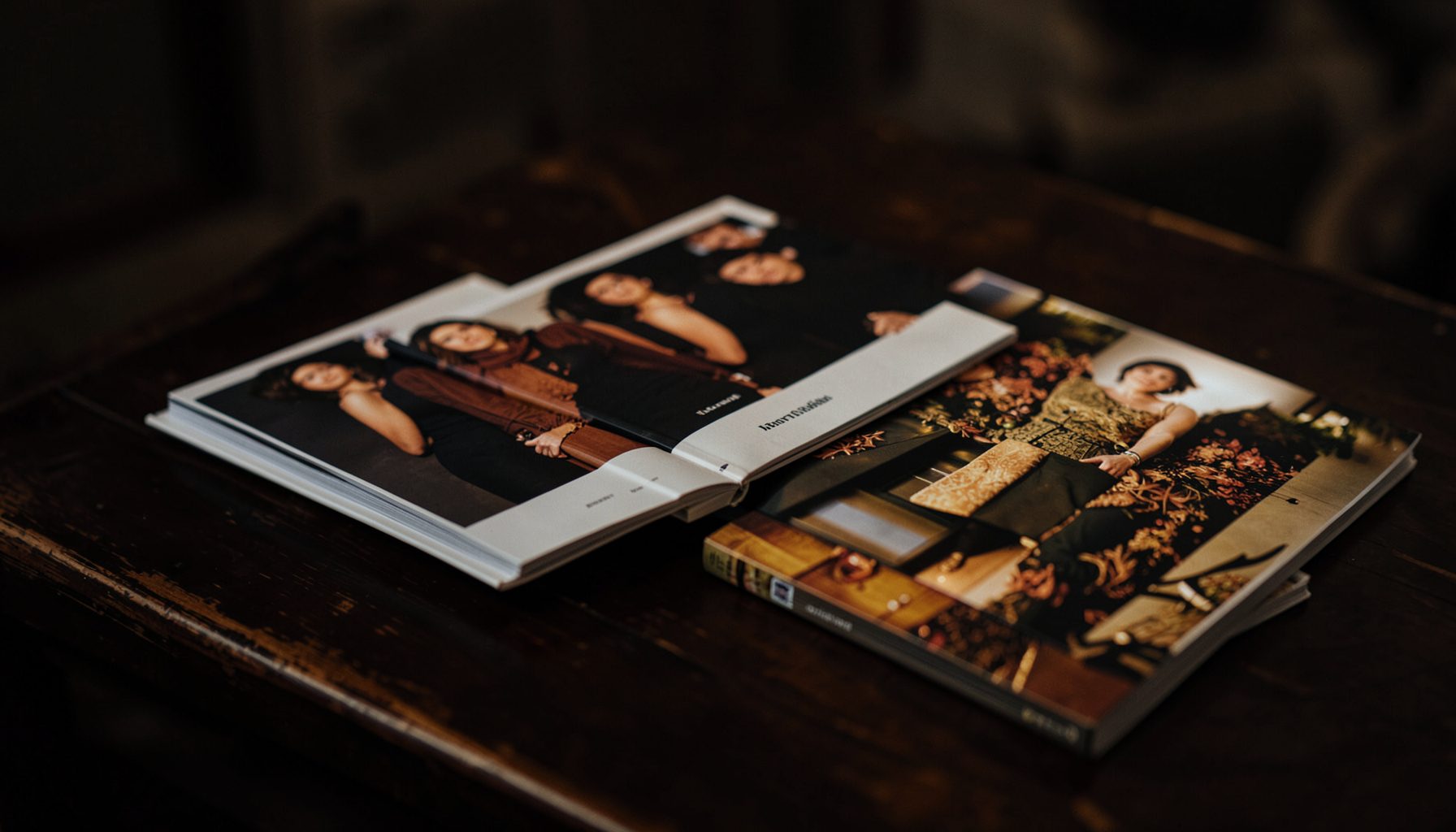 A moody still life of fashion monographs and glossy art books stacked on a dark wooden table, deep browns and warm golds, soft shadows